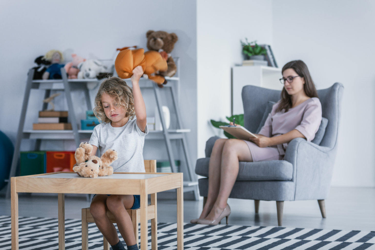 A child with behavioral problems sitting by a table and hitting a teddy bear during a therapeutic meeting with a therapist in a family support center.